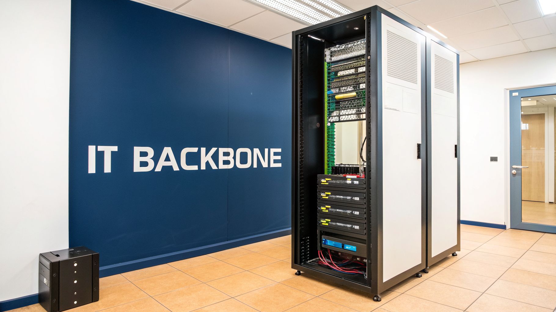 A server room with an open IT rack displaying hardware, next to a blue wall saying 'IT BACKBONE'.